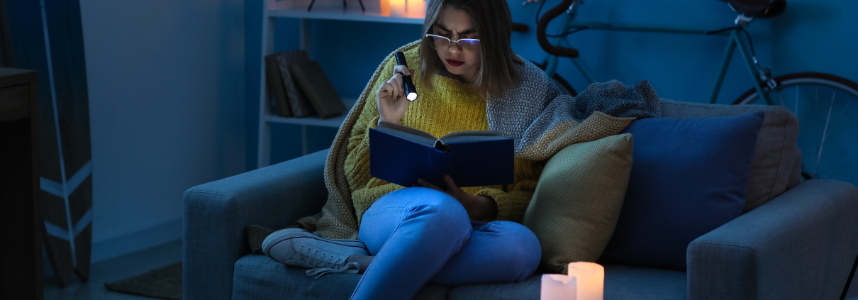 Woman sitting on a couch using a torch to read in a dark home during load shedding in South Africa, highlighting the need for a reliable inverter setup